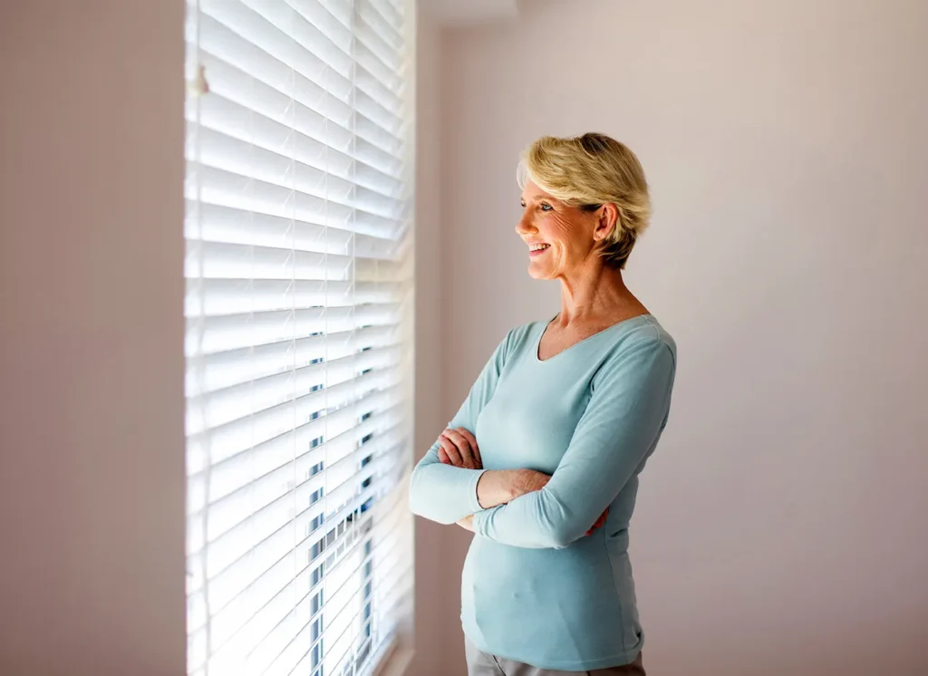 Woman standing beside a window fitted with white wooden Venetian blinds