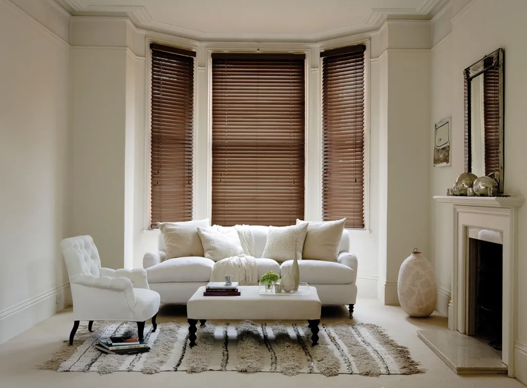 Dark walnut wooden Venetian blinds in a bay window above a cream sofa and fireplace
