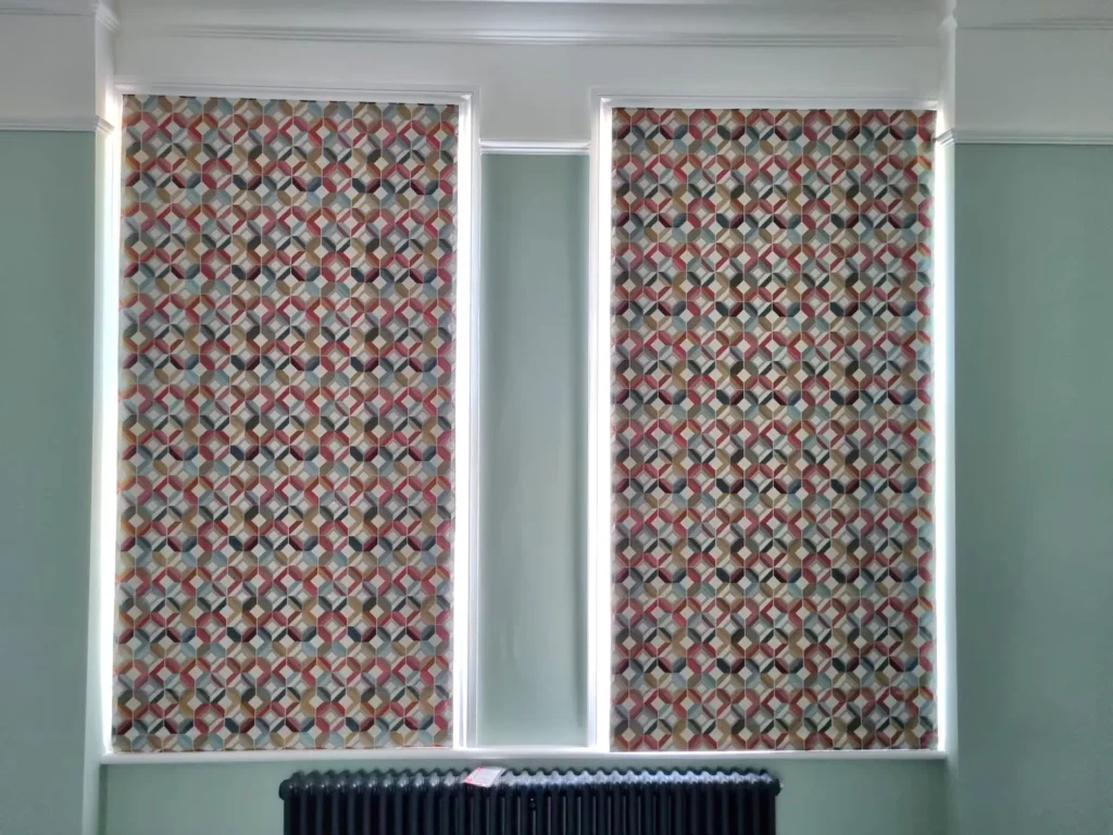 A pair of boldly patterned roller blinds fully lowered against a sage-green wall